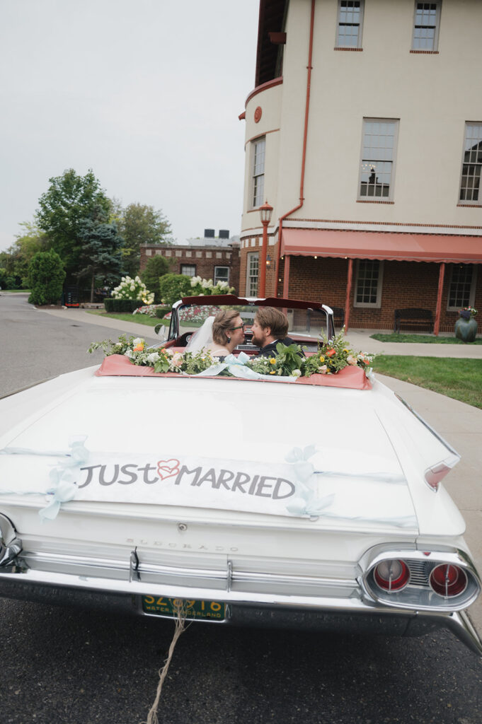 Bride and groom touching noses in a decked out vintage Eldorado with florals and a Just Married sign on the back outside of Detroit Yacht Club.