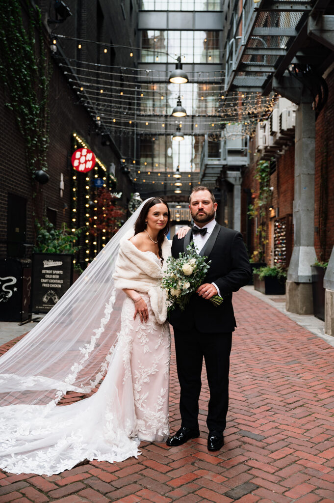 Bride and groom standing together in The Belt alley in downtown Detroit with string lights overhead.