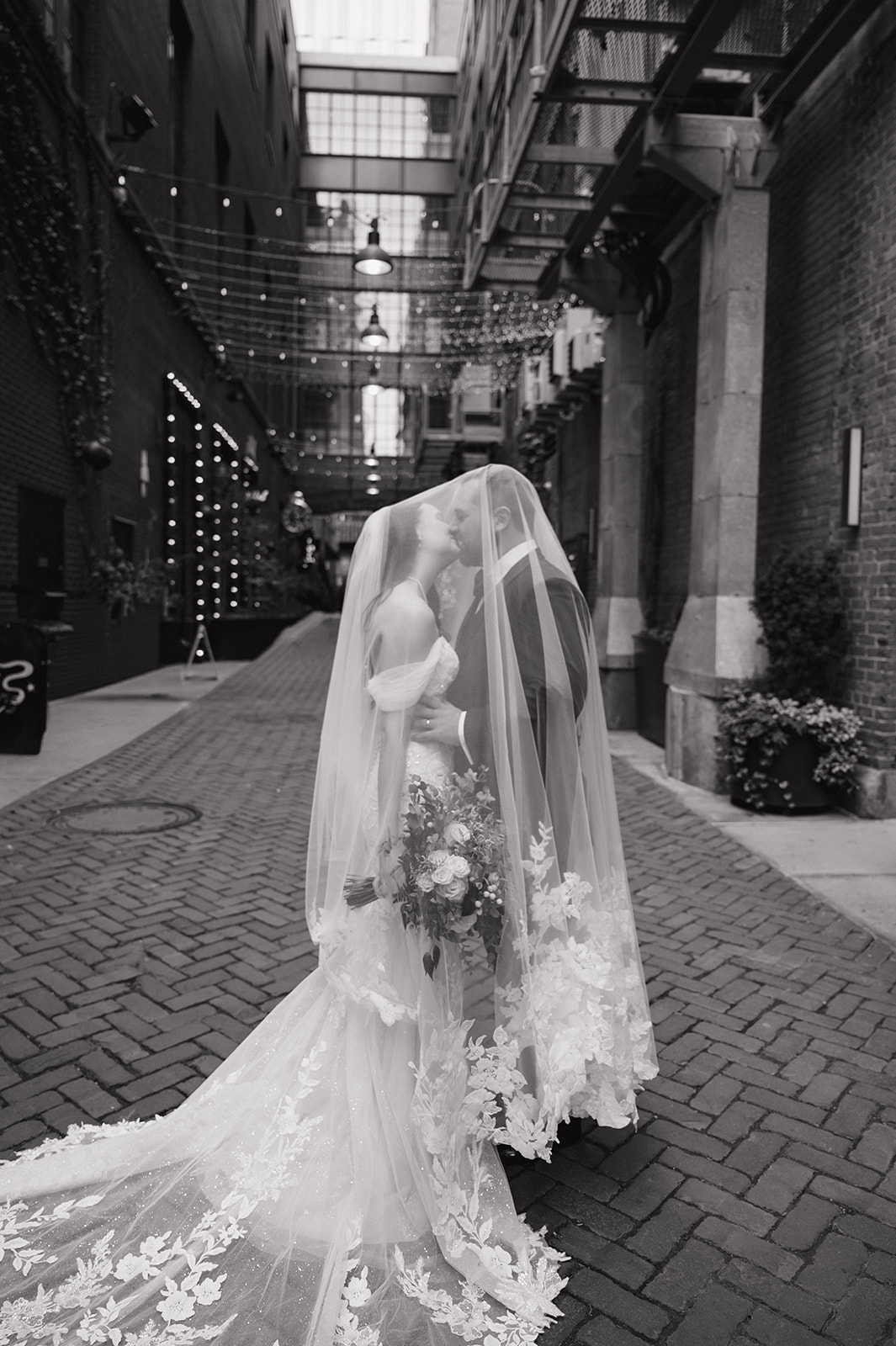 Black and white portrait of a bride and groom kissing under a veil in The Belt alley in downtown Detroit under string lights.