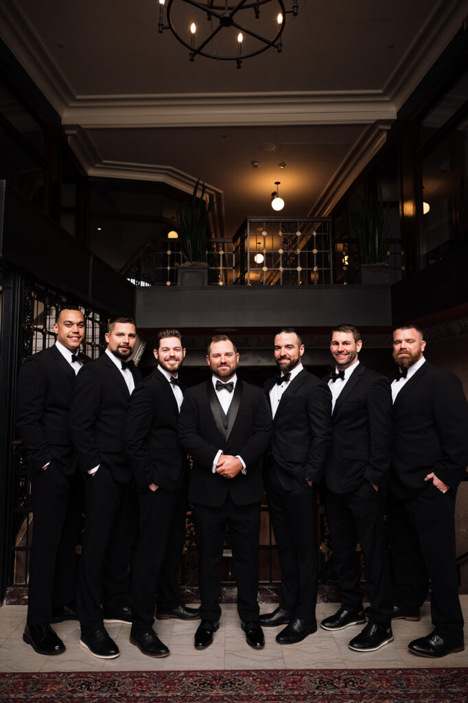 Groom and groomsmen posing together in black tuxes inside hotel during wedding portraits.
