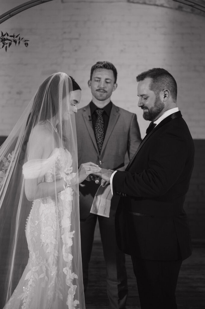 Black and white photo of a bride and groom exchanging rings during their ceremony.
