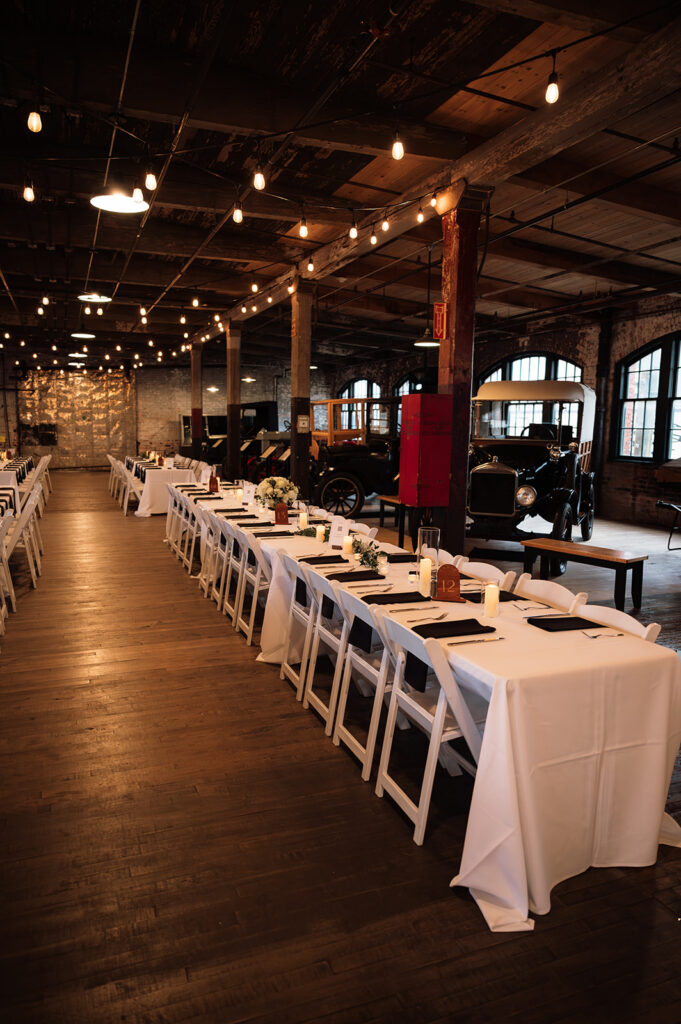Reception tables set with white linens and candles inside Ford Piquette Avenue Plant surrounded by vintage cars.