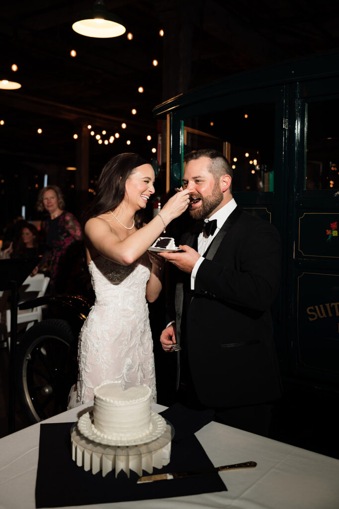 Bride feeding groom cake during reception under string lights at Ford Piquette Avenue Plant.