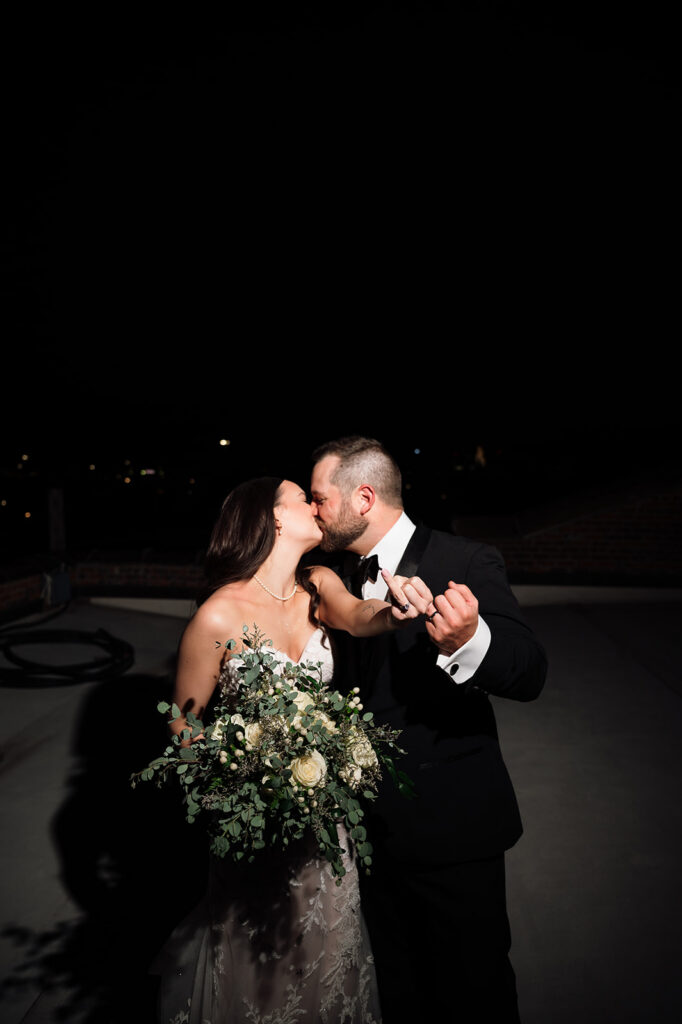 Bride and groom kissing and celebrating during nighttime wedding portraits with bouquet.