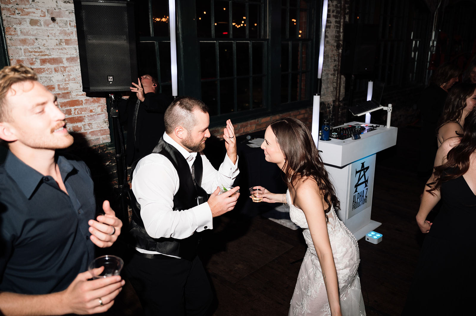 Bride and groom dancing together during reception with guests and DJ in background.