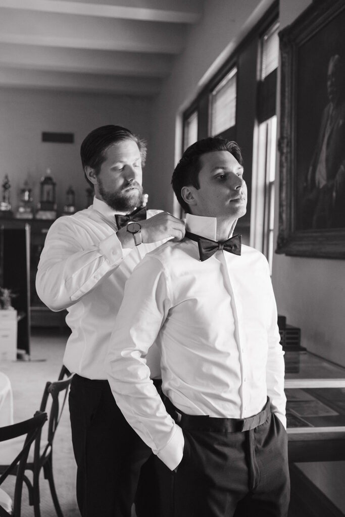 Black and white photo of a groom helping his groomsmen get dressed.