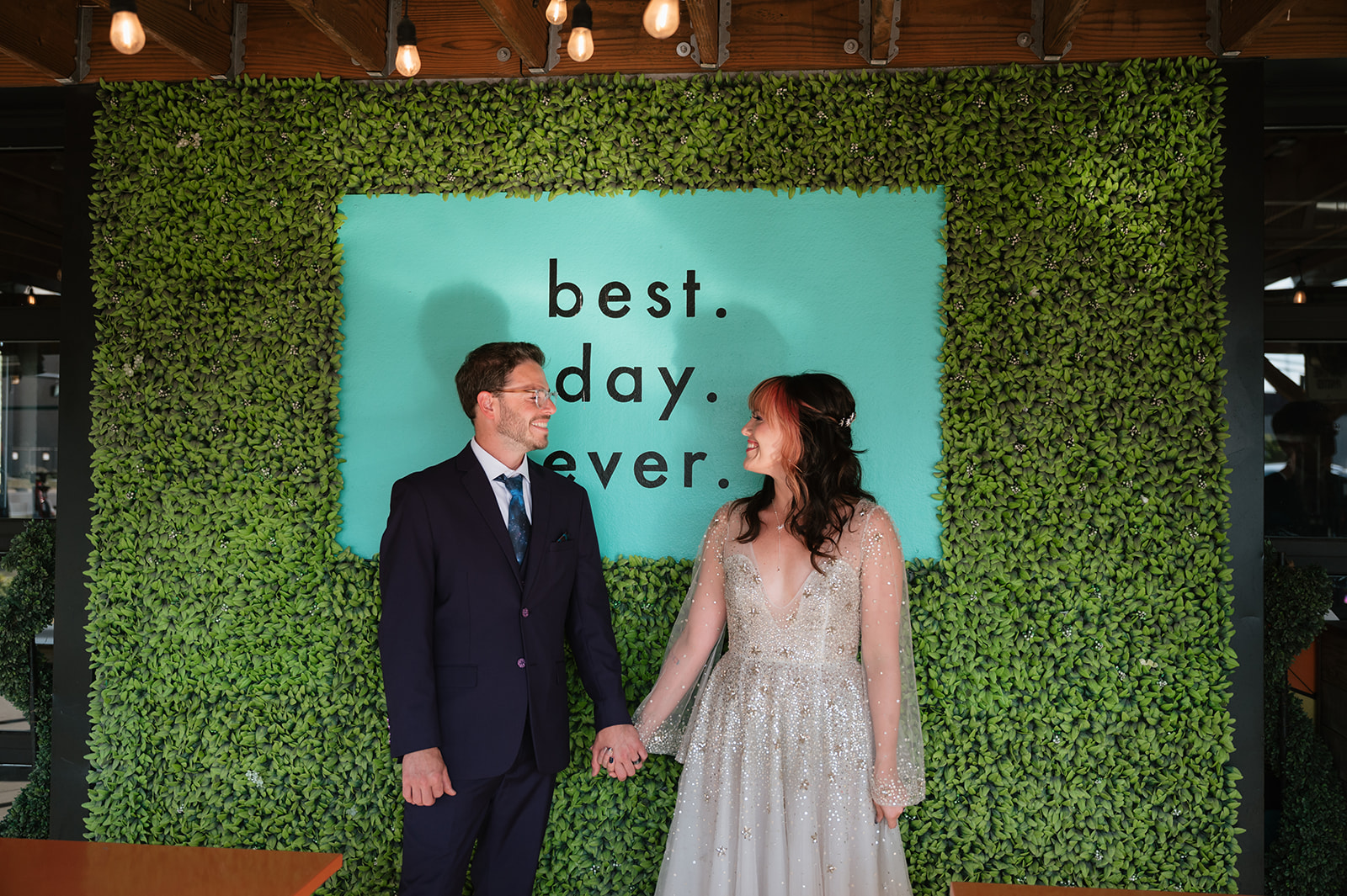 Bride and groom holding hands in front of a greenery wall at Detroit Fleat.