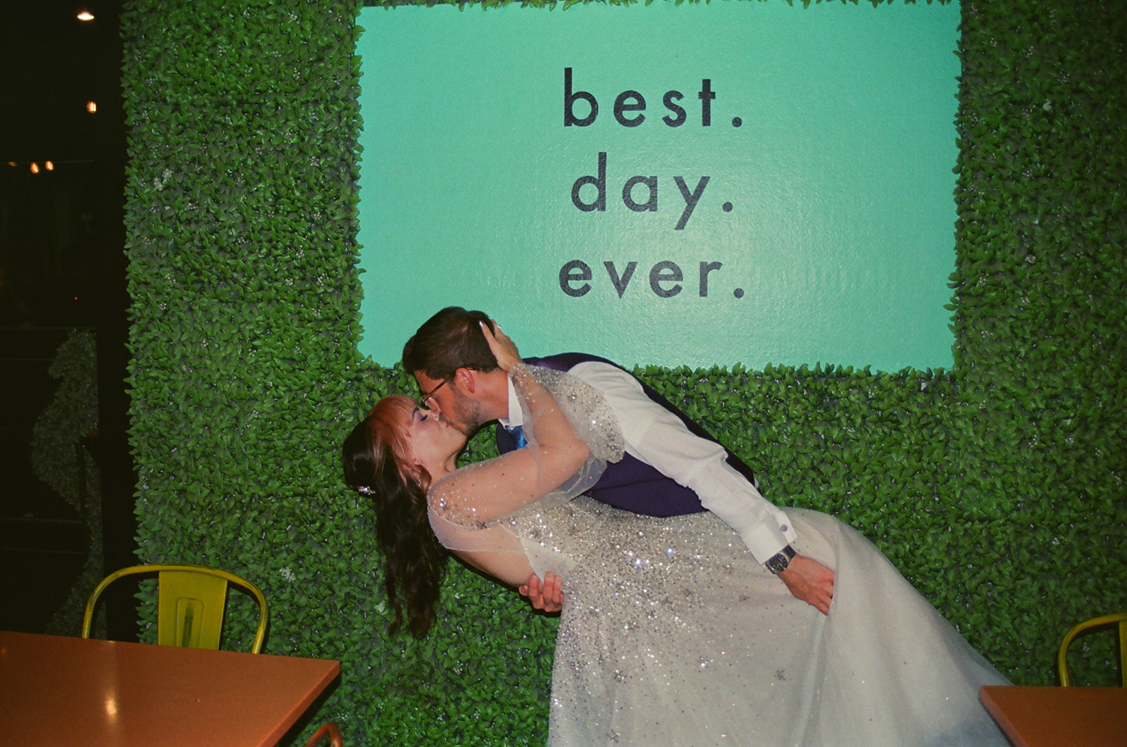 Bride and groom kissing in front of a greenery wall that reads best day ever on film.