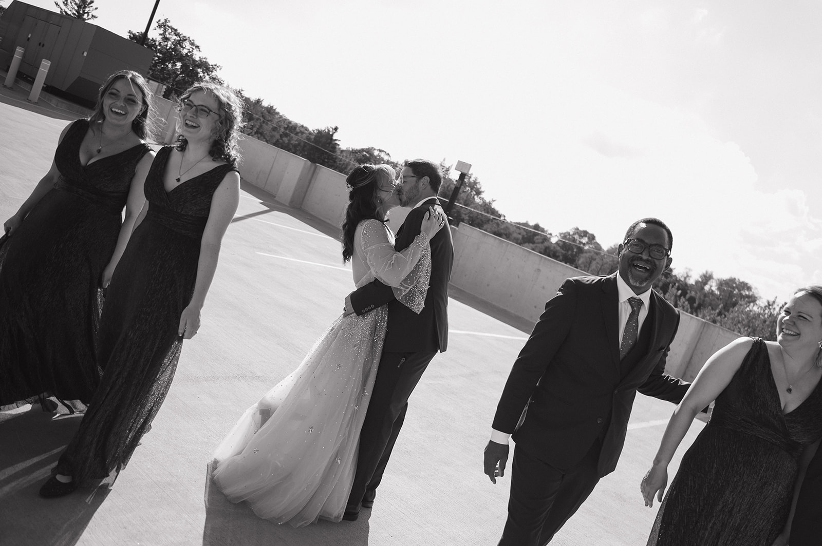 Black and white portrait of a bride and groom kissing surrounded by their wedding party on a rooftop.