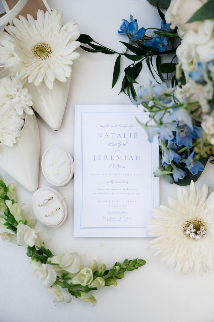 Flat lay of blue and white wedding details with flowers, invitation, shoes, and rings.