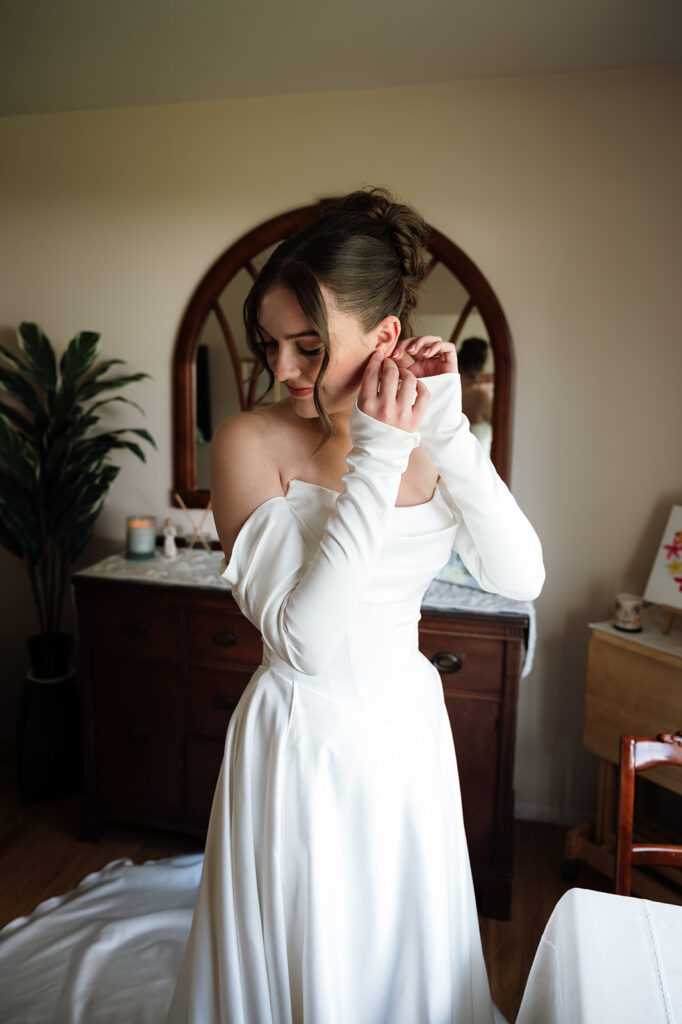 Bride putting on earrings in her off the shoulder wedding dress before the ceremony.