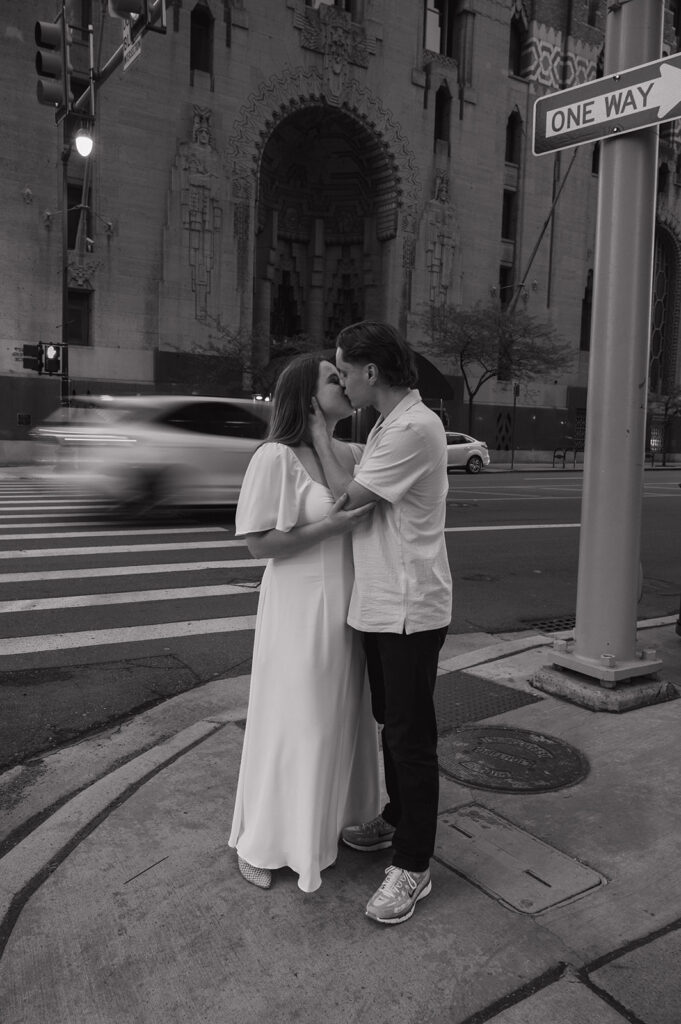 Couple kissing on a downtown Detroit sidewalk with a one way sign and city traffic behind them.