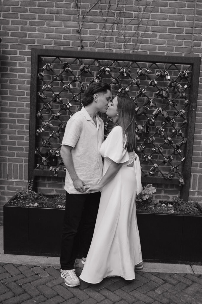 Couple kissing in front of a brick wall and lock installation in The Belt.