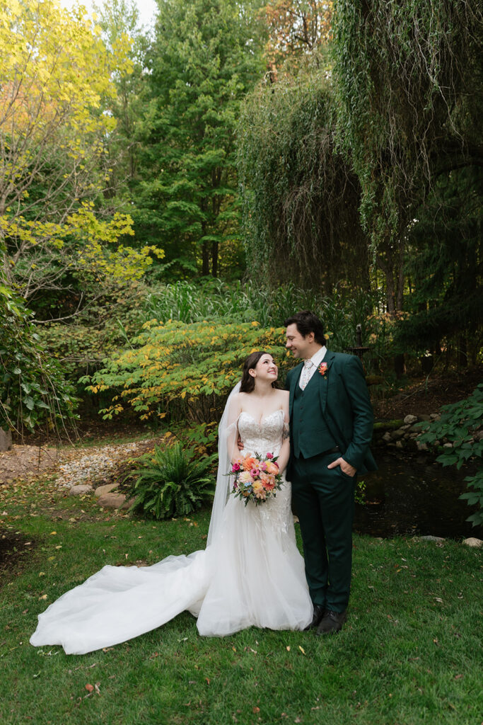 Bride and groom posing beneath a willow tree beside a garden pond in Owosso, Michigan at Everlastings in the Wildwood.