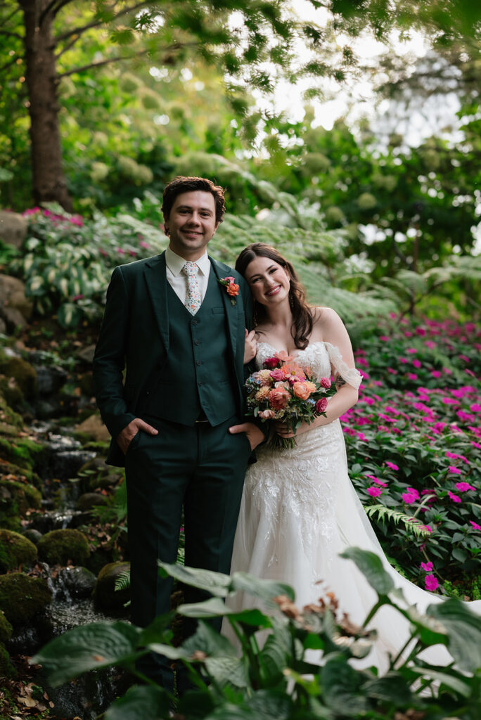 Bride and groom surrounded by ferns, colorful flowers, and a waterfall at Everlastings in the Wildwood.