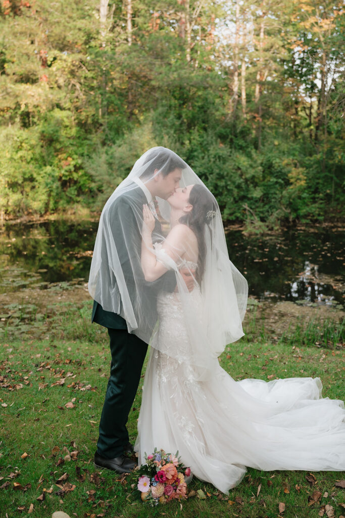 Bride and groom kissing beneath the bride’s veil beside a quiet garden pond.
