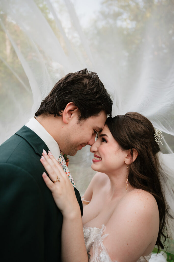 Bride and groom smiling at each other as they pose under the brides veil. 