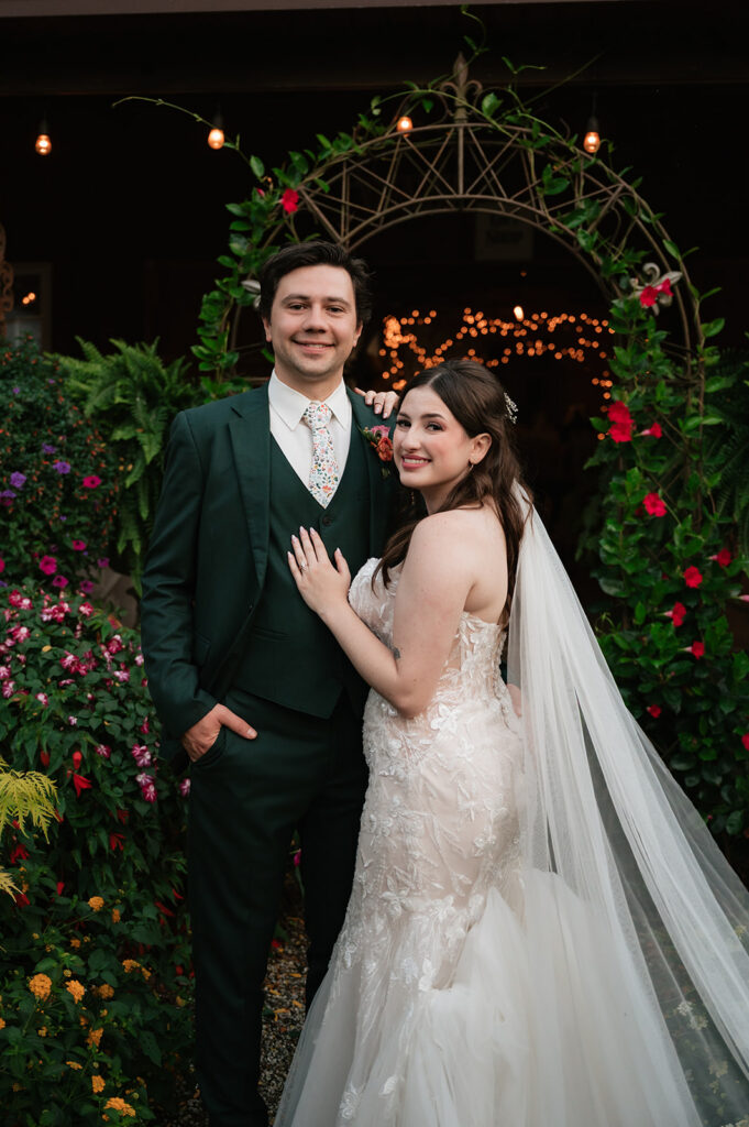 Bride and groom posing beneath a greenery-covered metal arch with string lights behind them at Everlastings in the Wildwood.