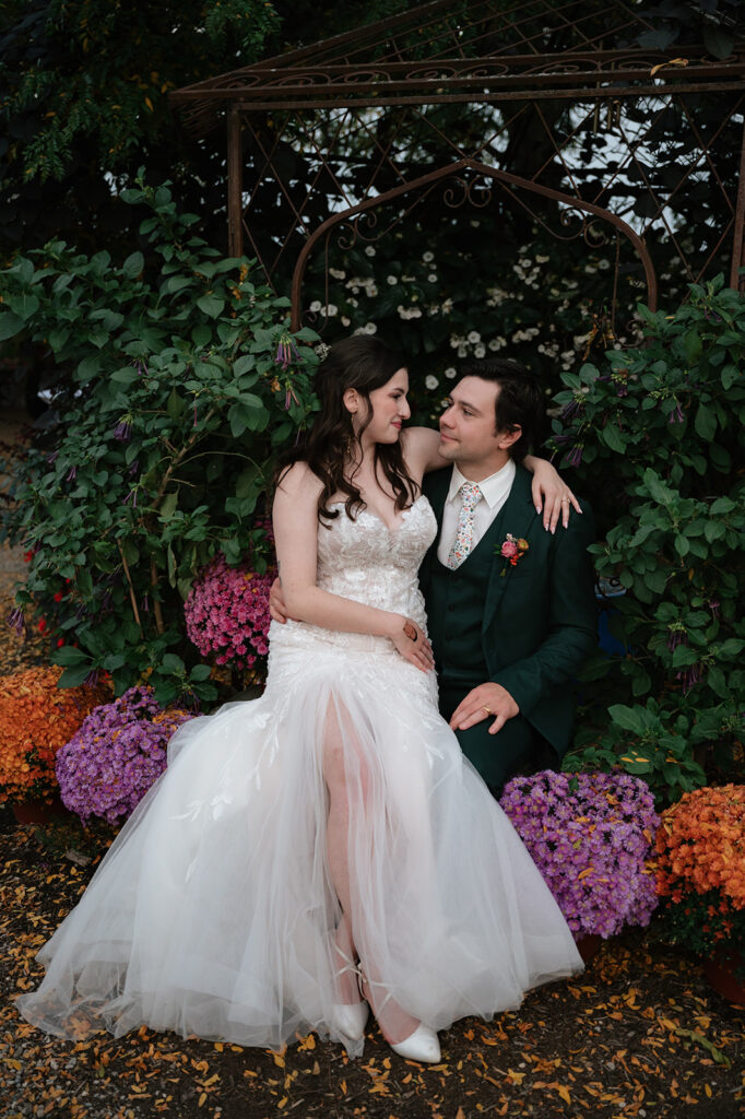 Bride and groom sitting beneath a wrought iron garden arch surrounded by colorful flowers at Everlastings in the Wildwood.