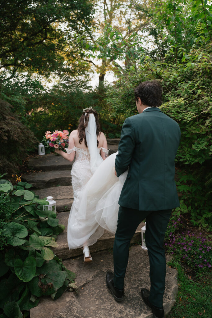 Groom carrying the bride’s train as they walk along a stone path through the gardens at Everlastings in the Wildwood.