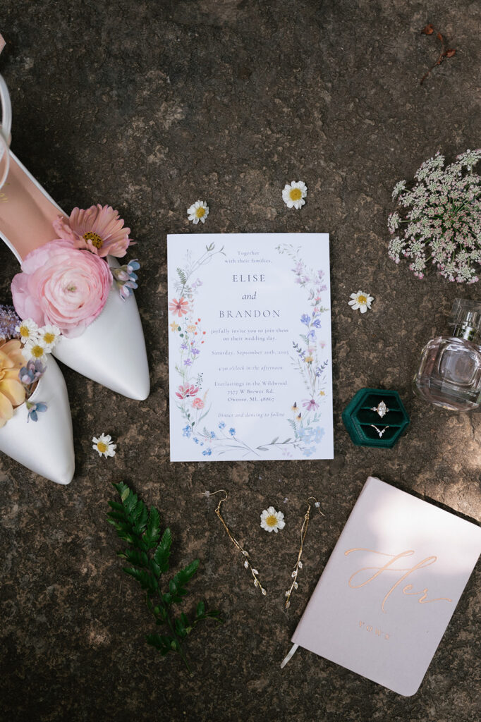 Floral wedding invitation flat lay with white shoes, rings, perfume, jewelry, and vow book.