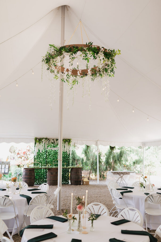 Tented reception space with white tables, dark green napkins, string lights, and a greenery chandelier from a Everlastings in the Wildwood wedding reception.