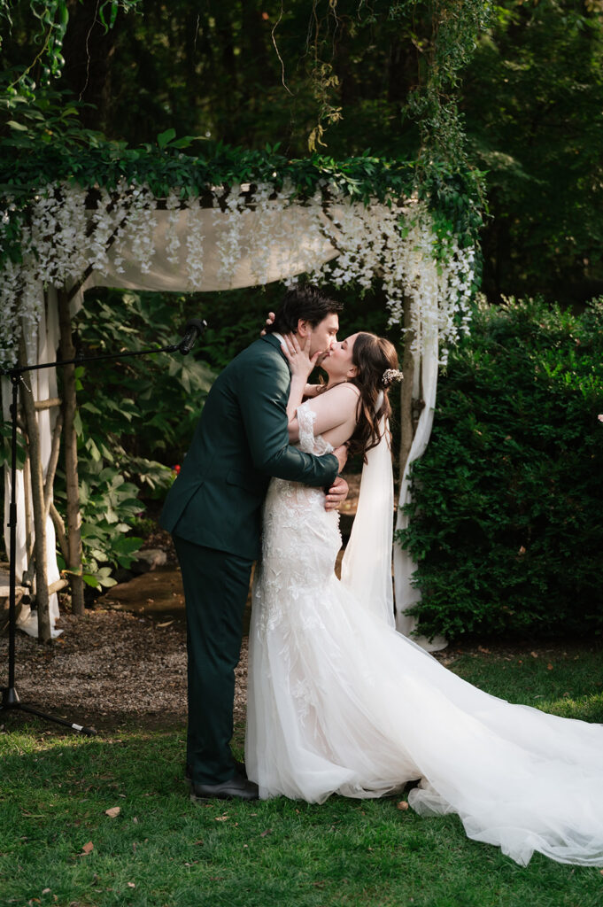 Bride and groom sharing their first kiss beneath a flower-covered garden arbor.