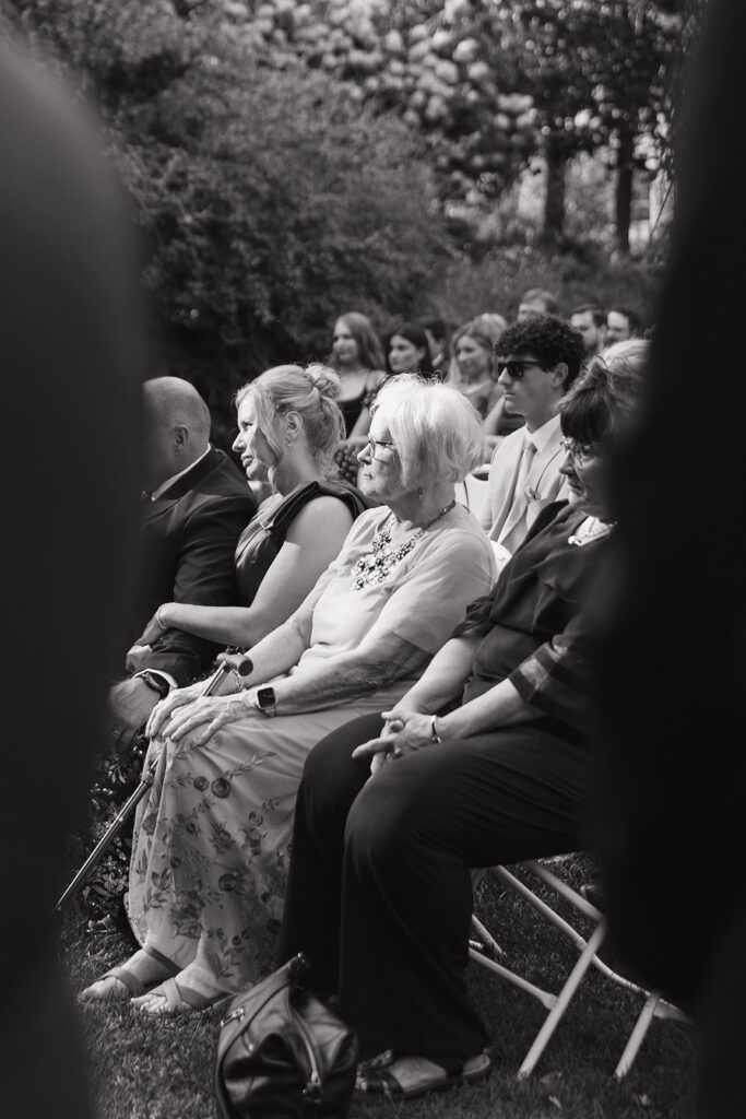 Black and white candid photo of guests watching an outdoor ceremony.