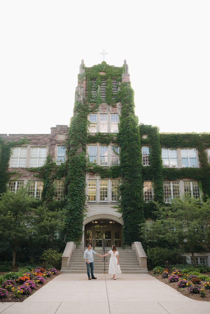 Couple holding hands in front of an ivy-covered building during Aquinas College engagement photos.