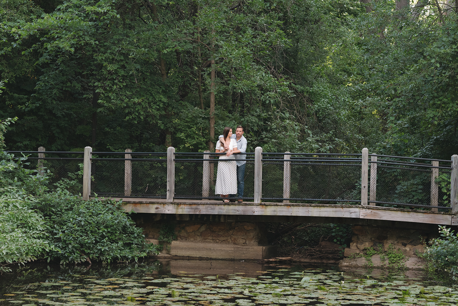 Couple embracing on a wooden bridge surrounded by trees at Aquinas College.