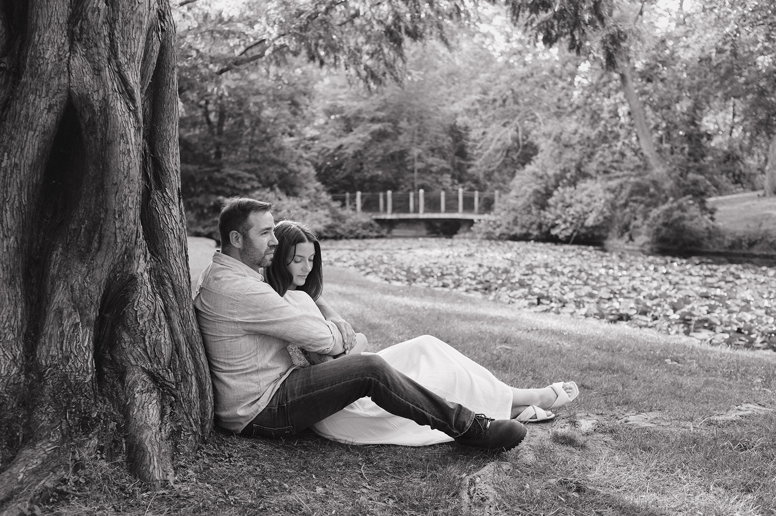 Black and white photo of a couple sitting against a tree beside a lily pond during their engagement session.