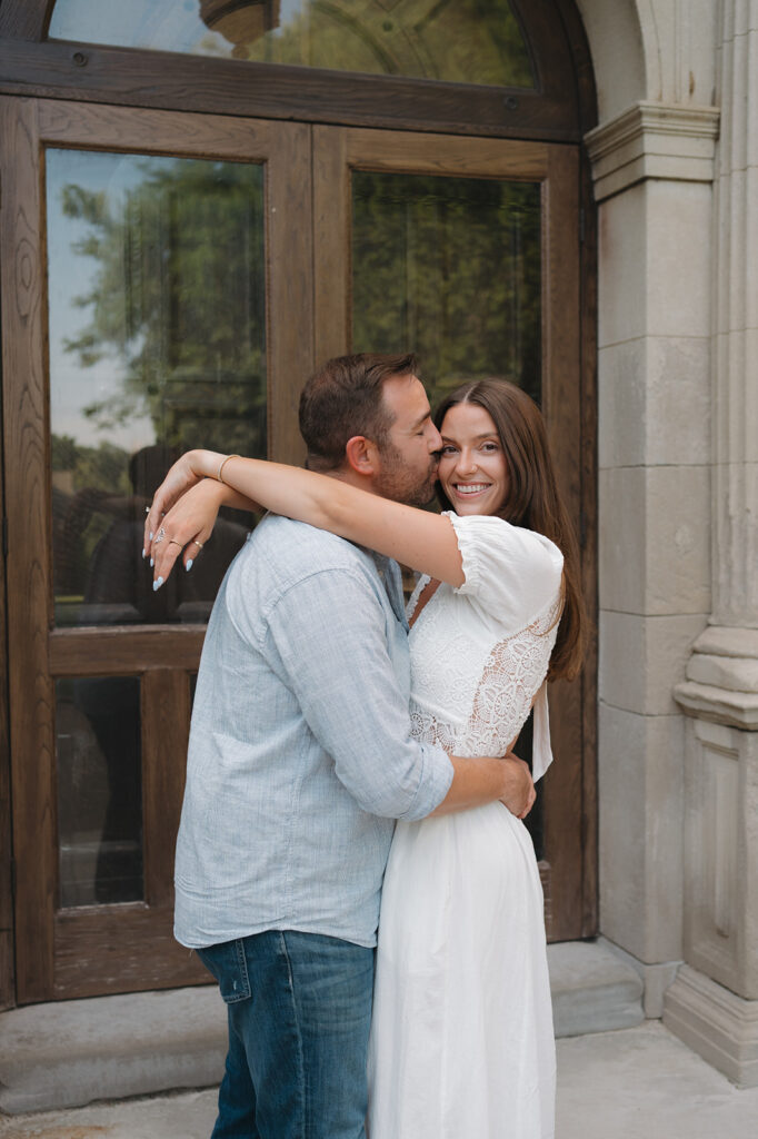 Man kissing his fiancé in front of an arched wooden doorway at Aquinas College.