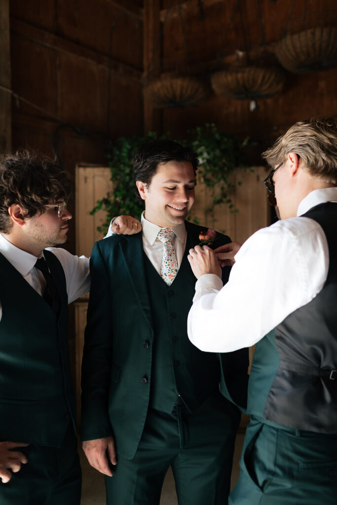 Groomsmen pinning a boutonniere onto the groom’s dark green suit while getting ready.