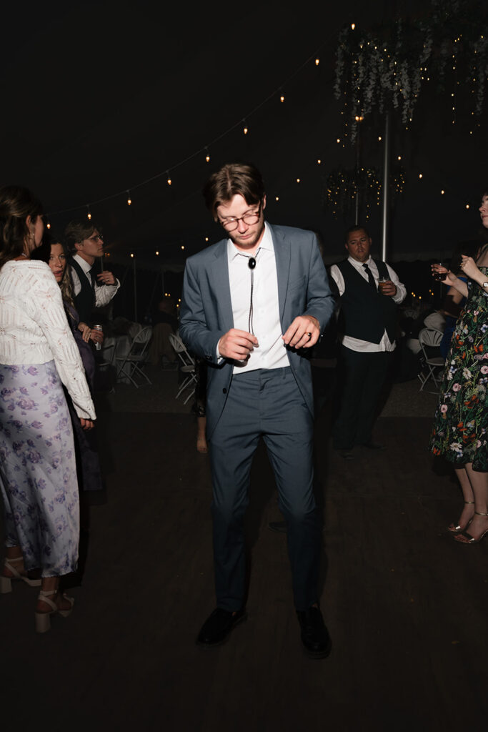 Wedding guest in a blue suit dancing beneath string lights inside the reception tent.