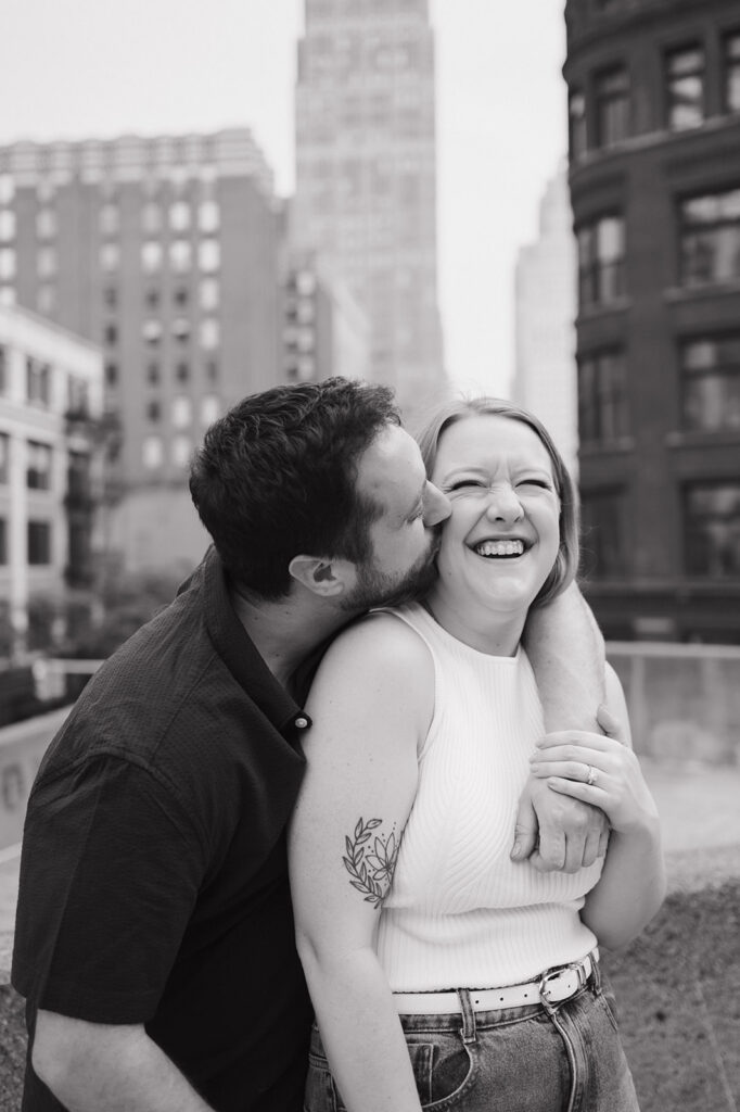 Man kissing his fiancés cheek during their rooftop Detroit engagement photos.