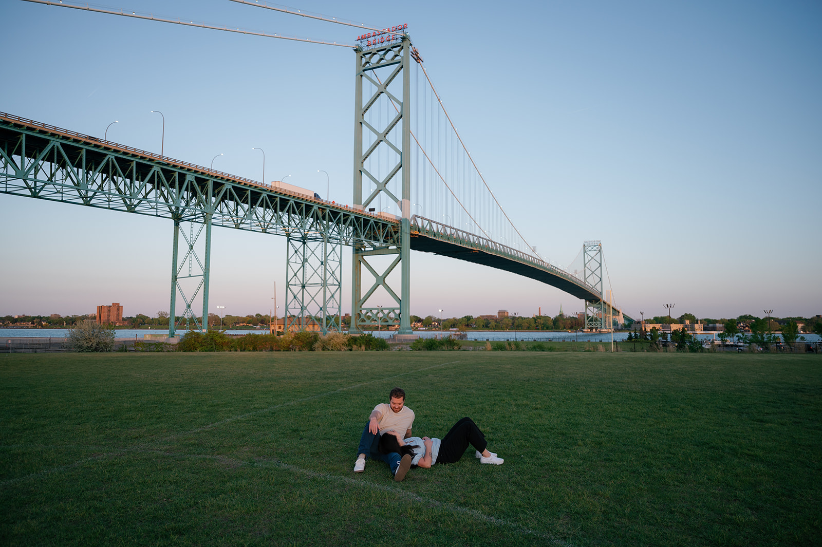 Couple laying in the grass for their Detroit engagement photos with Ambassador Bridge in the background. 