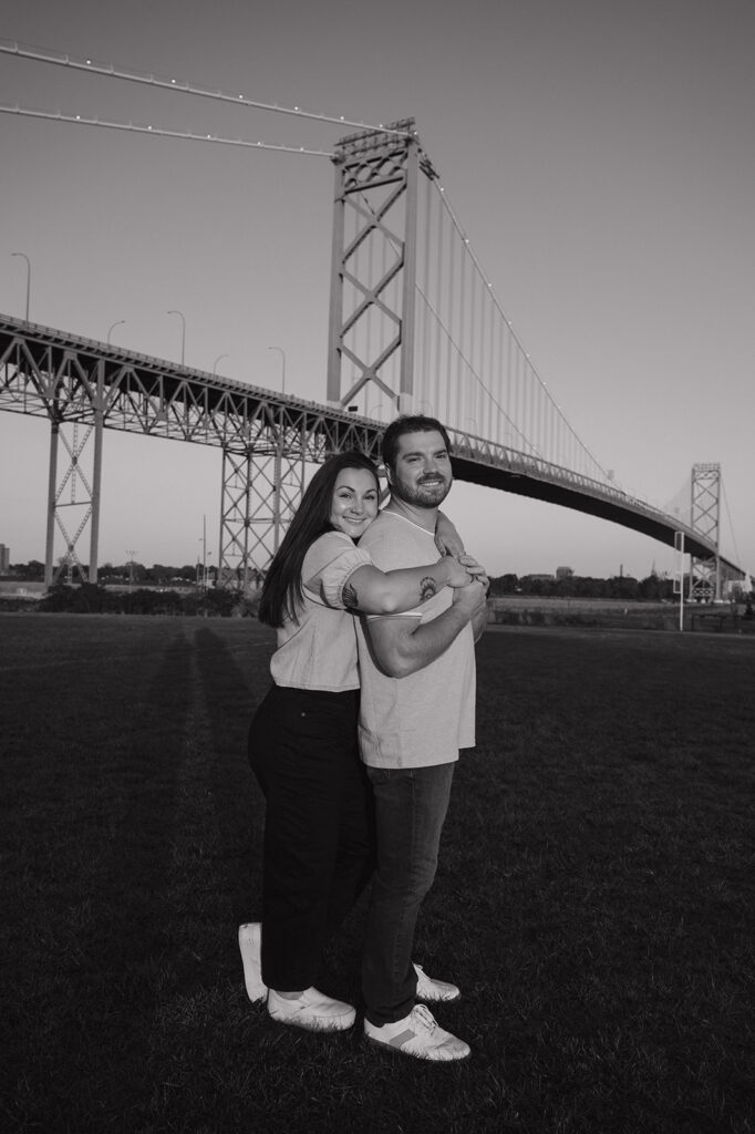 Black and white photo o a couple posing with Ambassador Bridge in the background. 