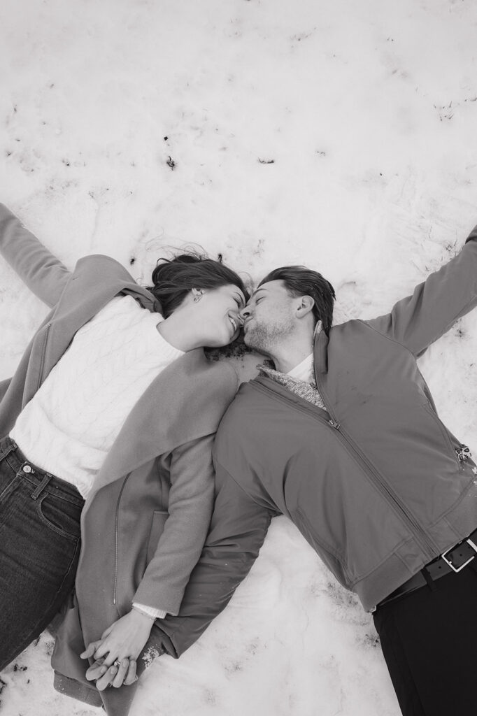 Couple laying and kissing in the snow during their winter engagement photos. 